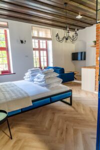 Łóżko z białą pościelą i stertą poduszek, niebieska sofa, drewniany strop, żyrandol i duże okna z roletami